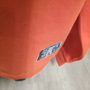 Op de afbeelding: Oranje sweatshirt met een blauwe en witte patch op de mouw. De patch heeft een grafisch ontwerp van een gestileerde figuur.