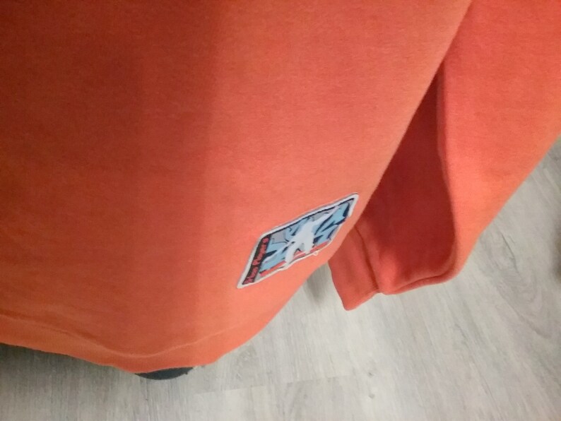 Op de afbeelding: Oranje sweatshirt met een blauwe en witte patch op de mouw. De patch heeft een grafisch ontwerp van een gestileerde figuur.