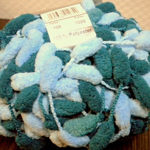 Może przedstawiać: Motka przędzy w kolorze turkusowym i jasnoniebieskim z pomponami. Przędza jest w 100% z poliestru. Etykieta zawiera napis "Farbe 105" i "1029".