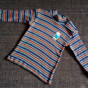 Op de afbeelding: Een langarmig shirt met blauwe, oranje en groene strepen en een klein cartoonfiguurtje op de voorkant.