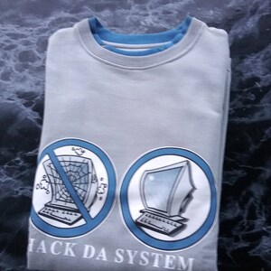 Può includere: Felpa grigia con una grafica blu e bianca di un computer con una ragnatela sullo schermo e il testo "Hack Da System".