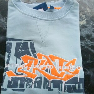 May include: A light blue sweatshirt with a graphic print of a stylized graffiti design in orange and white. The text "GETTING JUMPED IN THE" is visible on the print.