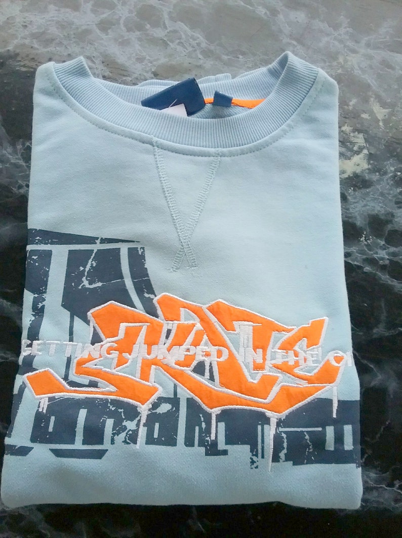 May include: A light blue sweatshirt with a graphic print of a stylized graffiti design in orange and white. The text "GETTING JUMPED IN THE" is visible on the print.