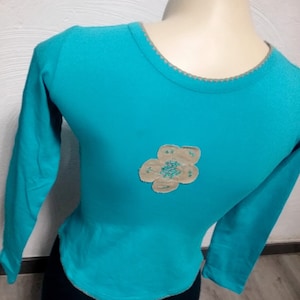 Könnte beinhalten: Ein türkisfarbenes Langarmshirt mit einem beigefarbenen Blumenapplikation auf der Vorderseite.