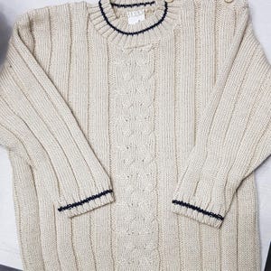 Puede incluir: Suéter de punto trenzado color crema con mangas y cuerpo acanalados. El suéter tiene un ribete azul marino en el cuello y los puños. Dos pequeños botones redondos de color claro están en el hombro. El suéter está hecho de un material suave y texturizado.
