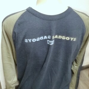 May include: A long-sleeved, navy blue t-shirt with a green and brown color block design on the sleeves. The shirt has a crew neck and features the text "BADBOYS" printed on the back in a faded, multi-colored font.