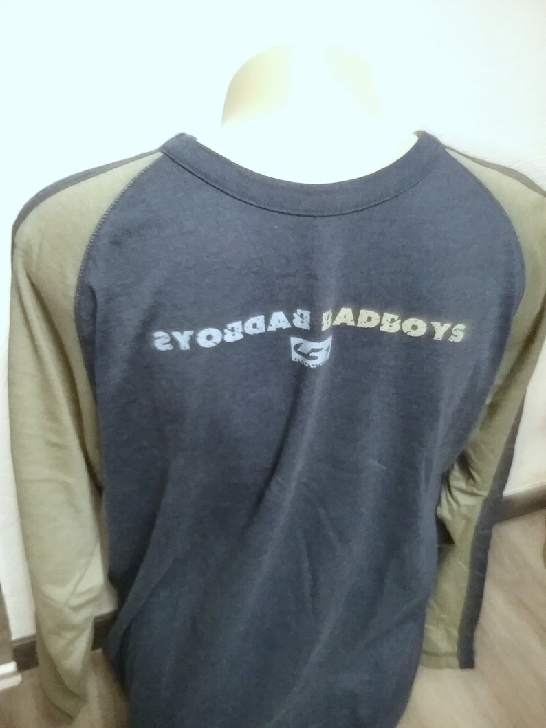 May include: A long-sleeved, navy blue t-shirt with a green and brown color block design on the sleeves. The shirt has a crew neck and features the text "BADBOYS" printed on the back in a faded, multi-colored font.