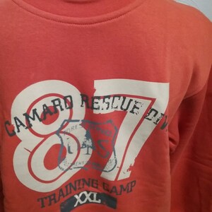 May include: Red sweatshirt with a white graphic print that says "Camard Rescue Div. 87" with a vintage style stamp design. The sweatshirt also says "Training Camp" and "XXL" in white.