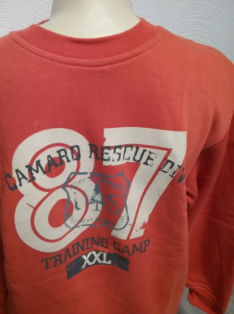 May include: Red sweatshirt with a white graphic print that says "Camard Rescue Div. 87" with a vintage style stamp design. The sweatshirt also says "Training Camp" and "XXL" in white.