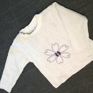 Può includere: Maglietta bianca a maniche lunghe con un piccolo fiore ricamato in viola e rosa sul davanti.