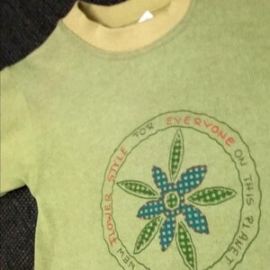Può includere: T-shirt verde chiaro con scollo tondo e maniche corte. La maglietta presenta una grafica circolare con un disegno floreale blu e verde, circondata dal testo "Flower Style, The New, This Is Planet, For Everyone On".