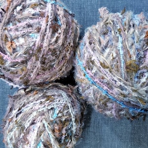Op de afbeelding: Drie bollen getextureerd garen in tinten bruin, grijs, roze en blauw. Het garen heeft een noppen, multi-vezel constructie, ideaal voor brei- of haakprojecten. De bollen zijn in een driehoekig patroon gerangschikt.