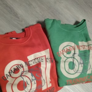 May include: Two sweatshirts, one red and one green, with the text "Camaro Rescue" and the number "87" printed on them. Both sweatshirts have a distressed, vintage look.