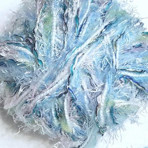 Könnte beinhalten: Ein Knäuel strukturierten Garns in Blau-, Weiß- und Türkistönen. Das Garn hat ein flauschiges, flauschiges Aussehen mit unterschiedlich dicken Strähnen. Die Farbpalette erzeugt eine weiche, luftige Ästhetik, die sich für Strick- oder Häkelprojekte eignet.