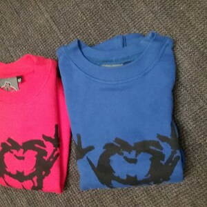 Könnte beinhalten: Zwei gefaltete Sweatshirts, eines rot und eines blau, mit einem schwarzen grafischen Design in Form eines Herzens auf jedem.