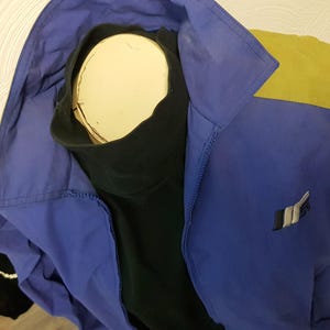 May include: A blue and yellow jacket with a black turtleneck. The jacket has a zipper and a logo on the front. The jacket is displayed on a mannequin.
