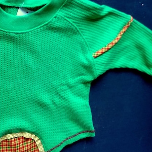 May include: Green waffle knit long-sleeved shirt with a red and yellow plaid trim on the sleeve and a red and yellow plaid trim on the bottom hem.