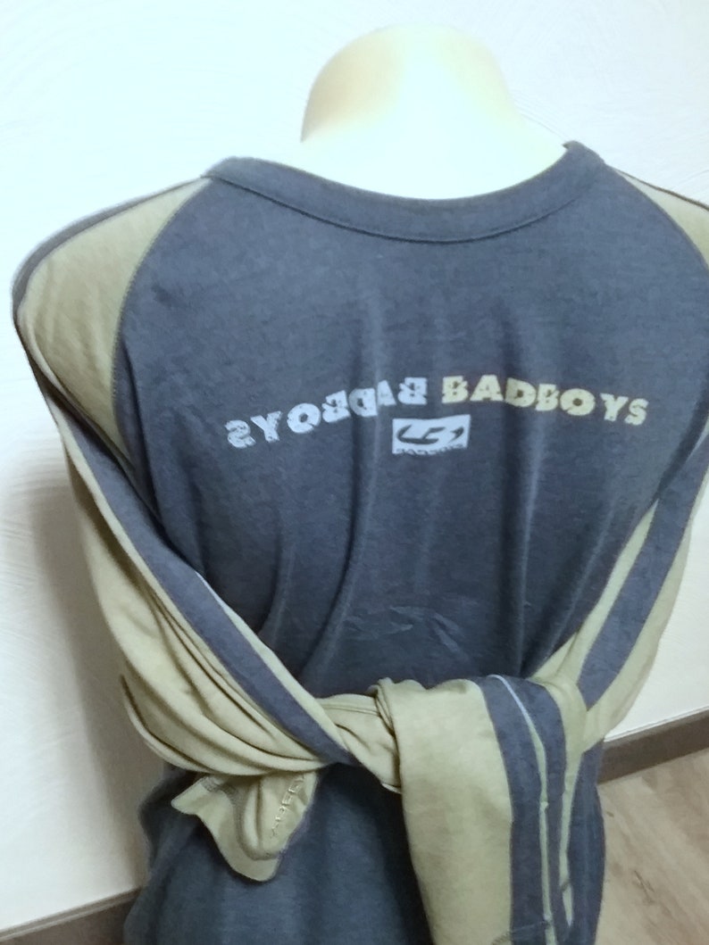 May include: A long-sleeved shirt with a gray body and tan sleeves. The shirt has a graphic print on the back that says "Badboys" with a logo underneath. The sleeves have a stripe design.