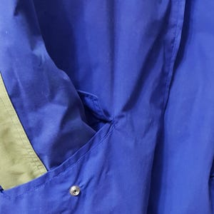 May include: Close-up of a blue jacket with a pocket and a snap closure. The jacket has a green accent on the side. The fabric appears to be a lightweight material, suitable for layering.