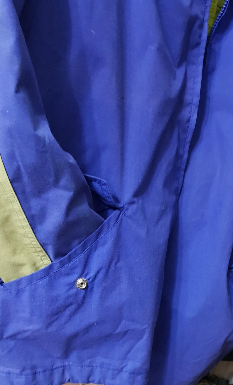 May include: Close-up of a blue jacket with a pocket and a snap closure. The jacket has a green accent on the side. The fabric appears to be a lightweight material, suitable for layering.