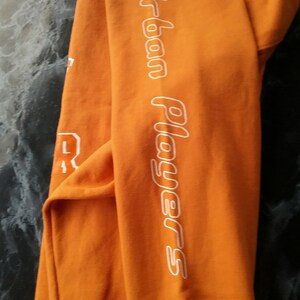 Op de afbeelding: Oranje sweatshirt met witte tekst "Urban Players" op de mouw.