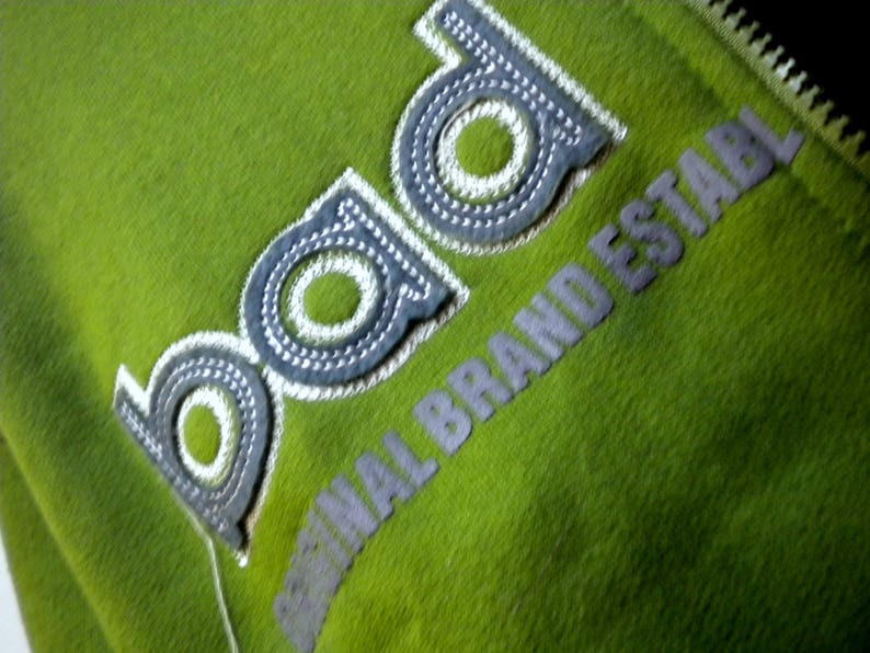 May include: A green zippered jacket with a white embroidered logo that says "bad original brand establ"