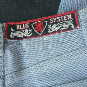 Puede incluir: Un par de pantalones vaqueros de color azul claro con un parche negro y rojo que dice "BLUE SYSTEM". El parche presenta un león y dos dragones.