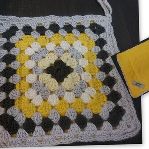 May include: A crocheted granny square cushion with a yellow, white, gray, and black color scheme. The cushion has a loop for hanging and a small yellow fabric tag with the text "Made with love" on it.