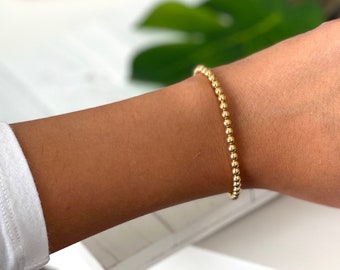 Puede incluir: Una pulsera dorada con cuentas se lleva en la muñeca. La pulsera está hecha de pequeñas cuentas redondas doradas ensartadas. El tono de piel es medio y la persona lleva una camisa blanca de manga larga.