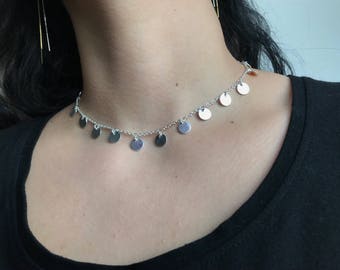 May include: A silver-colored choker necklace featuring small, round, flat disc pendants. The necklace is worn around the neck and is displayed against a black top. The necklace is a delicate piece of jewelry.