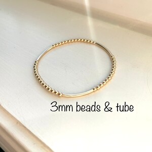 Op de afbeelding: Een gouden armband met kleine, ronde kralen en een buisvormige sluiting. 3mm beads & tube.