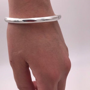 May include: A silver bangle bracelet worn on a person's wrist. The bracelet is plain and has a smooth, polished finish.