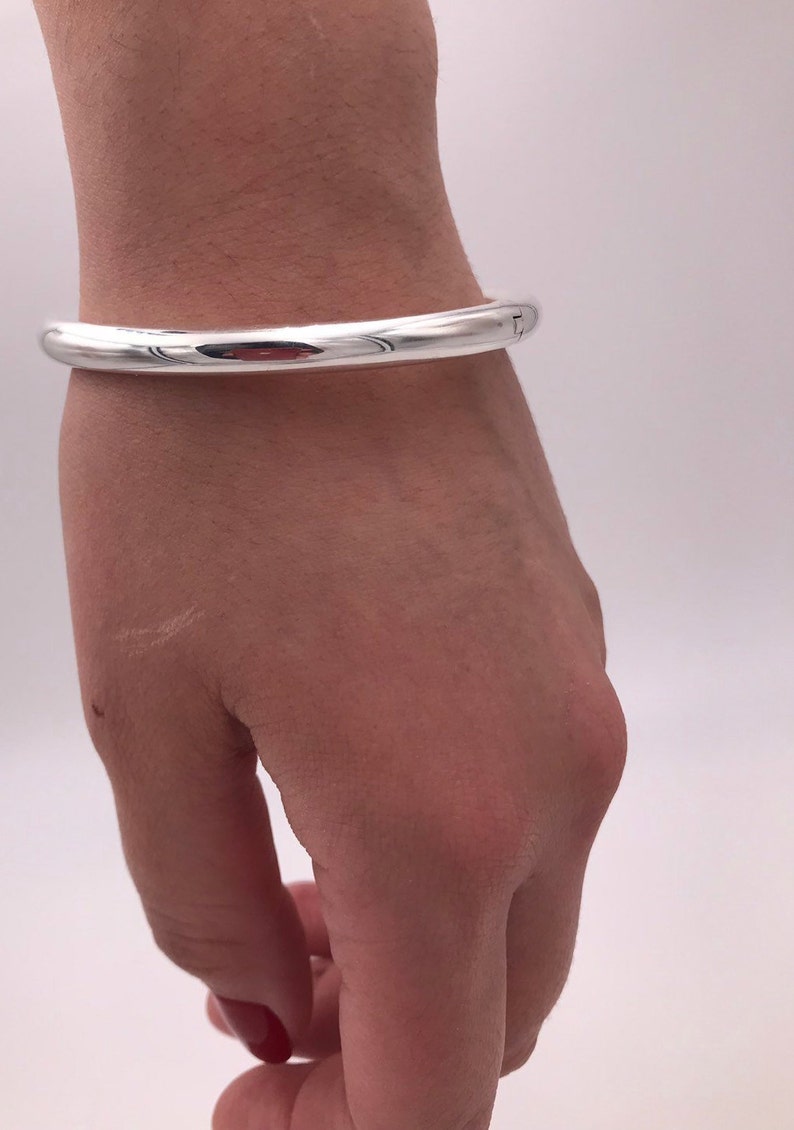 May include: A silver bangle bracelet worn on a person's wrist. The bracelet is plain and has a smooth, polished finish.