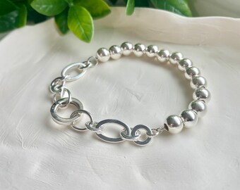 Puede incluir: Una pulsera de plata con una cadena de círculos entrelazados y una fila de cuentas de plata.