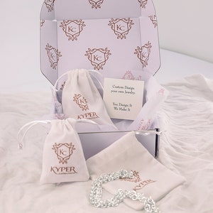 May include: A white box containing a silver chain necklace. The box is decorated with a pattern of the letters "KC" within a floral design. The box also includes a white drawstring bag with the text "KYPER collections" printed on it. The text "Custom Design your own Jewelry. You Design It We Make It" is printed on a white card inside the box.