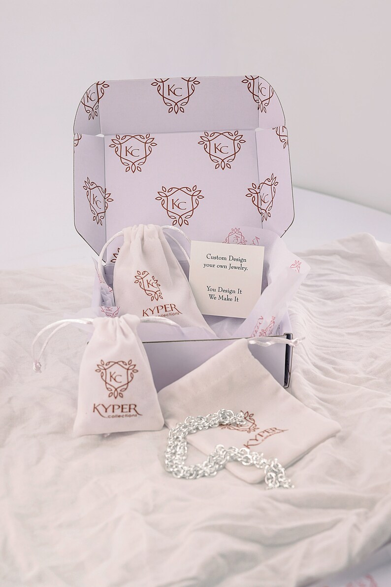 May include: A white box containing a silver chain necklace. The box is decorated with a pattern of the letters "KC" within a floral design. The box also includes a white drawstring bag with the text "KYPER collections" printed on it. The text "Custom Design your own Jewelry. You Design It We Make It" is printed on a white card inside the box.