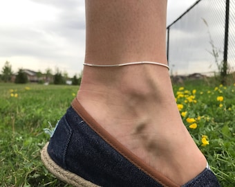 Puede incluir: Un tobillo de plata en el tobillo de una persona. El tobillo es una cadena fina con un diseño delicado. La persona lleva un zapato azul y marrón.