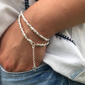 Könnte beinhalten: Ein doppelreihiges silbernes Armband mit facettierten Perlen. Das Armband hat eine verstellbare Kette mit einem kleinen Herzanhänger. Der Schmuck wird am Handgelenk getragen, mit einem Jeanshemd und Jeans im Hintergrund.