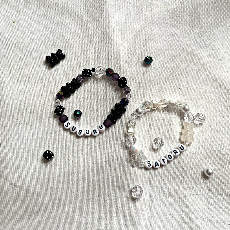May include: Two beaded bracelets with the names "SUGURU" and "SATORU" spelled out in white letters. The "SUGURU" bracelet is black with iridescent beads, while the "SATORU" bracelet is white with clear and iridescent beads.