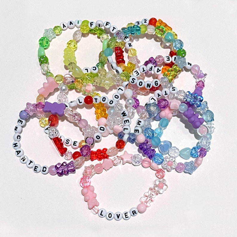 May include: A collection of colourful beaded bracelets with different words spelled out on them, including "Lover", "Enchanted", "We Were Happy", "22", "Ivy", "Fifteen", "All Too Well", "State of Grace", "You Are In Love", "Red", "Speak Now", "Fearless", and "1989".