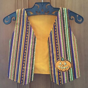 May include: A Halloween-themed vest with a colorful striped pattern in orange, purple, green, and black. The vest has a solid orange lining and a pumpkin applique on the front. It is hanging on a black hanger.