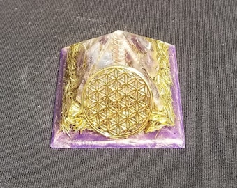 May include: A clear quartz pyramid with a gold flower of life symbol on top, surrounded by copper wire and purple resin. The pyramid is sitting on a dark gray surface.