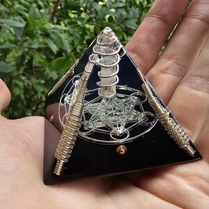 May include: Black obsidian pyramid with a clear quartz crystal point and copper wire wrapped around it. The pyramid has a geometric design inside.