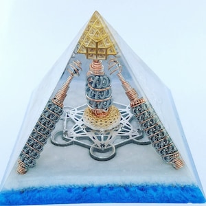 May include: A clear crystal pyramid with a gold-colored geometric design on the top. Inside the pyramid are three copper and silver wire coils, a silver metal flower of life design, and a gold-colored orb. The base of the pyramid is blue.