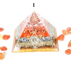 May include: A layered pyramid-shaped object with a clear resin exterior. The pyramid features red, silver, and gold layers, with a geometric design on the front. Surrounding the pyramid are scattered orange-red stones. The number "1" is visible at the top.