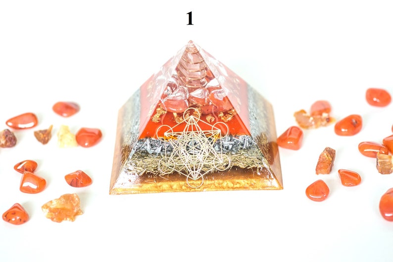 May include: A layered pyramid-shaped object with a clear resin exterior. The pyramid features red, silver, and gold layers, with a geometric design on the front. Surrounding the pyramid are scattered orange-red stones. The number "1" is visible at the top.