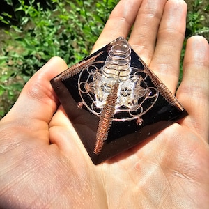 May include: A black obsidian pyramid with a clear quartz crystal inside. The pyramid is adorned with copper wire and a geometric design.