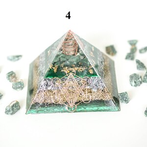 May include: A green pyramid-shaped orgone energy generator with gold accents and a copper coil at the apex. The pyramid features a geometric design and is surrounded by small green stones. The number "4" is visible at the top.