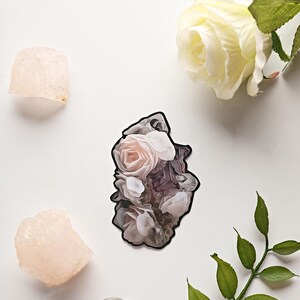 May include: A sticker of a rose bouquet in pink, white, and purple, with a black outline. Two pink crystals, a clear sphere, and a white rose with green leaves are arranged on a white surface.