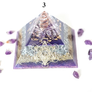 May include: A purple and silver pyramid-shaped orgone energy generator with a copper coil at the top. The pyramid features a Metatron's Cube design and is surrounded by amethyst crystals. The number "3" is visible at the top.